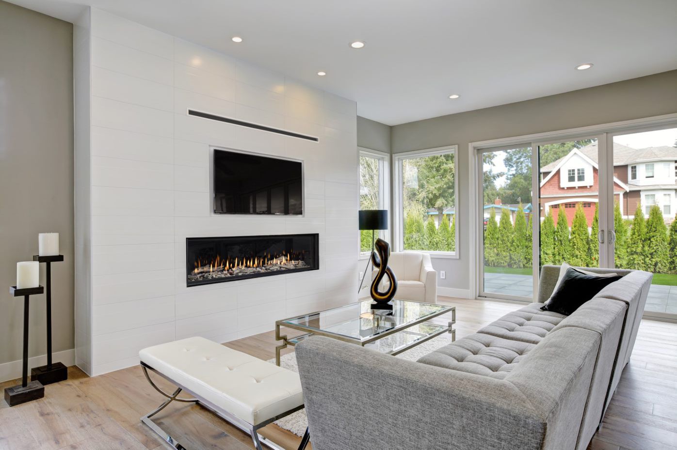A modern living space with a black, white, and gray color scheme, light wooden floors, floor-to-ceiling windows, and a large feature wall with a linear gas fireplace and a flatscreen TV.
